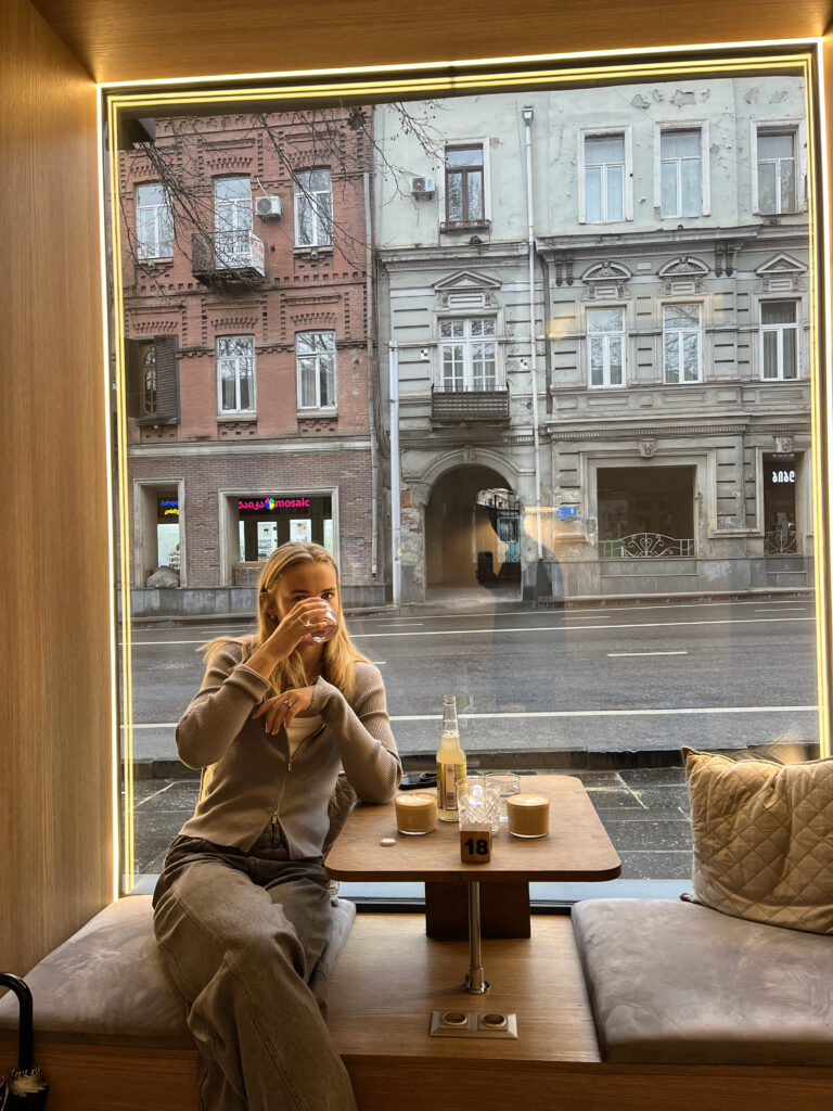 Ung kvinde sidder i et stort vinduesparti på en hyggelig café i Tbilisi og drikker kaffe med udsigt til klassiske georgiske bygninger