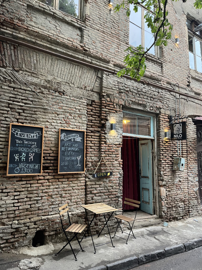 Rustik murstensfacade på caféen NILO med kridttavler for events og et lille udendørs cafébord med stole i Tbilisis gamle bydel