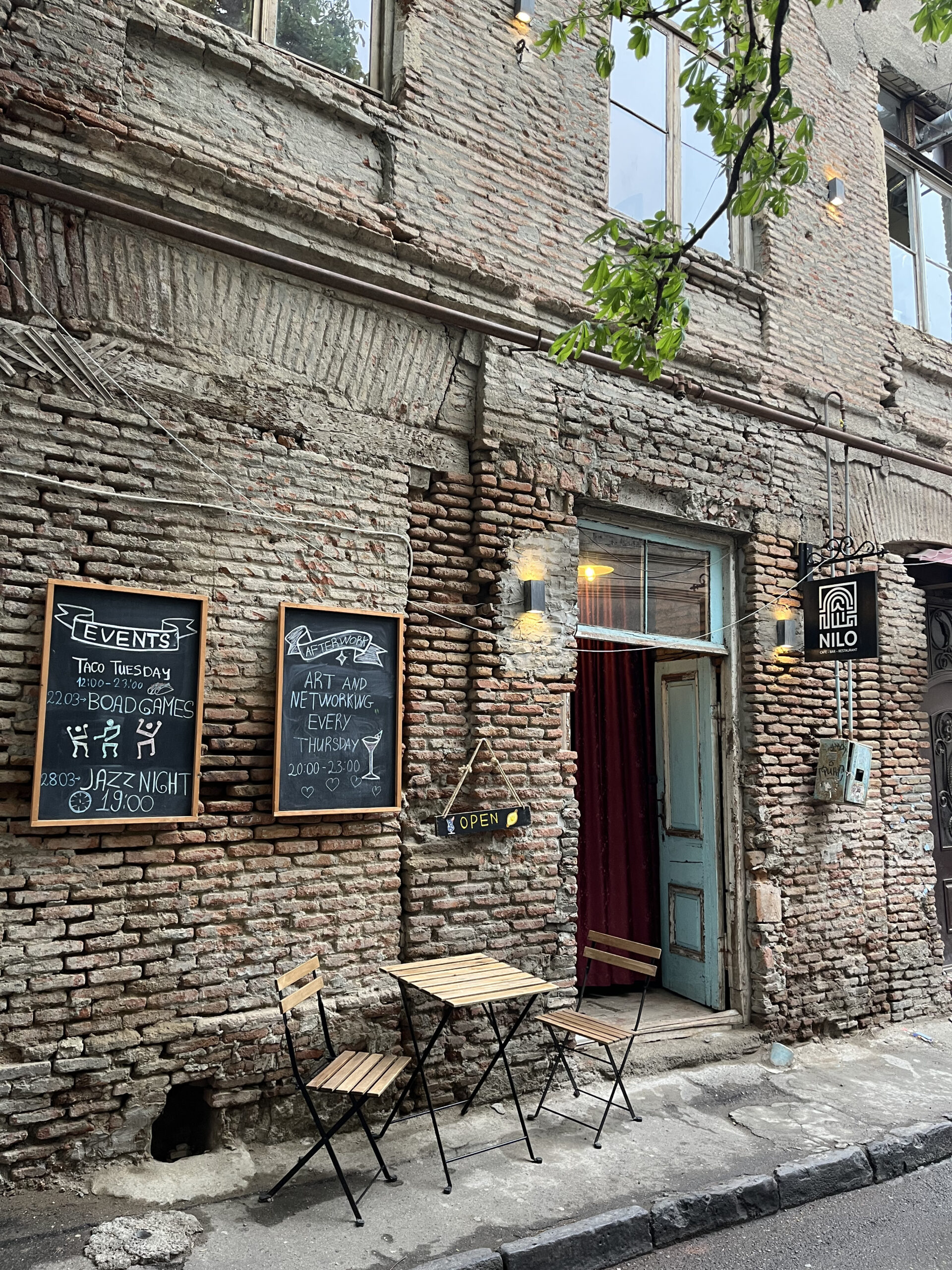 Rustik murstensfacade på caféen NILO med kridttavler for events og et lille udendørs cafébord med stole i Tbilisis gamle bydel