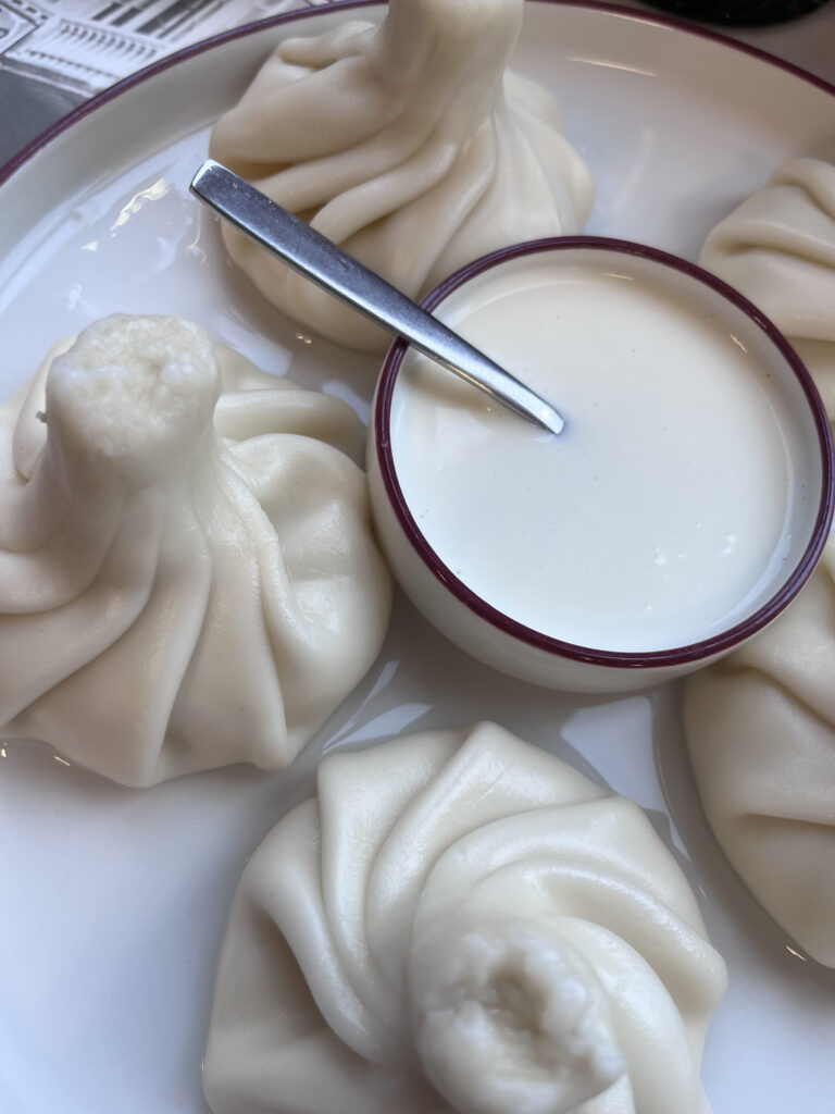 Tætbillede af traditionelle georgiske khinkali-dumplings serveret på en hvid tallerken med en skål cremet dip og en ske