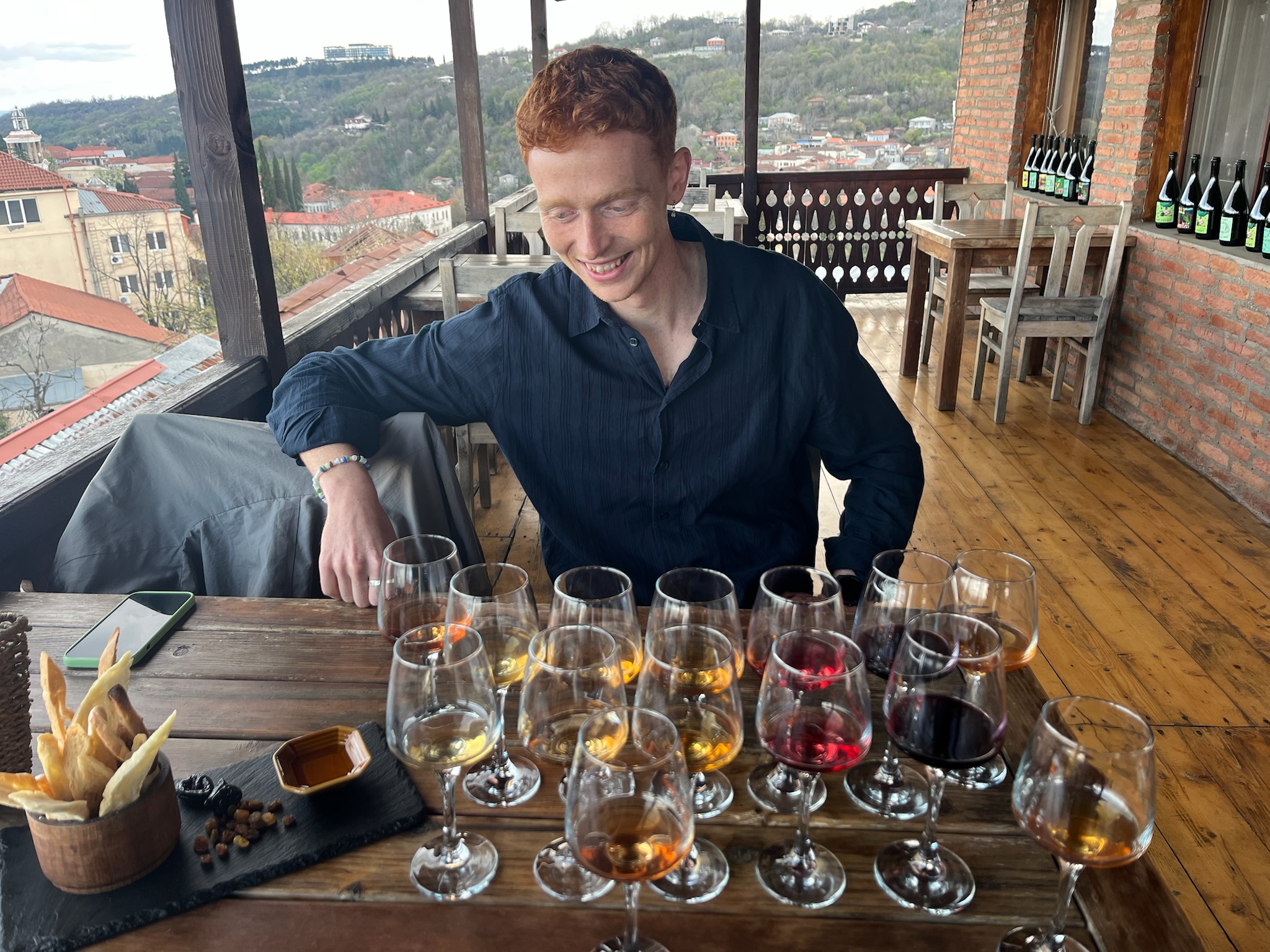 Ung mand smiler ved et træbord fyldt med adskillige glas rød- og ravfarvet vin til en klassisk georgisk vinsmagning med udsigt over byen