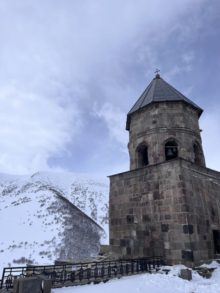 Historisk klokketårn i sten ved den berømte Gergeti Treenighedskirke mod en sneklædt bjergside i Kazbegi