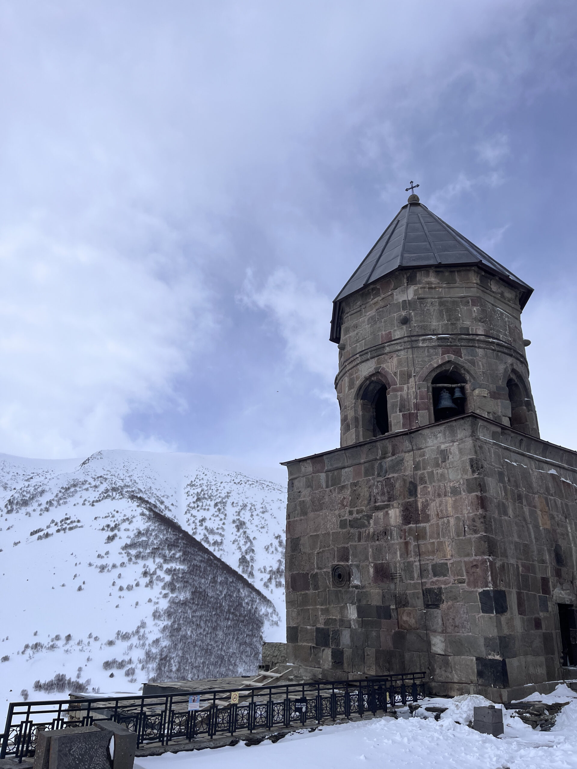 Historisk klokketårn i sten ved den berømte Gergeti Treenighedskirke mod en sneklædt bjergside i Kazbegi