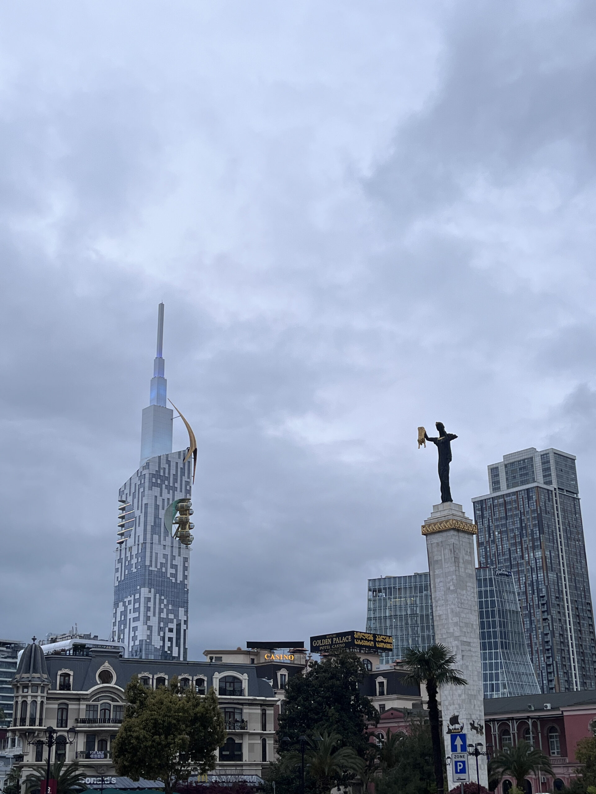 Medea-statuen på en høj søjle i Batumi med futuristiske skyskrabere og moderne arkitektur i baggrunden under en overskyet sky