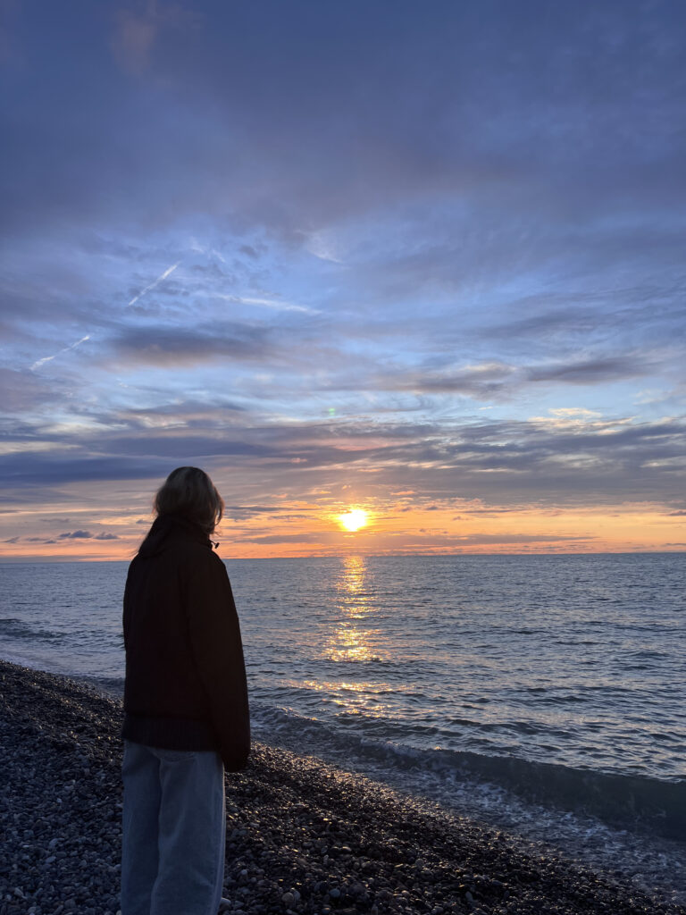 En kvinde ses fra ryggen, mens hun kigger ud over Det Sorte Hav på en stenstrand under en gylden solnedgang