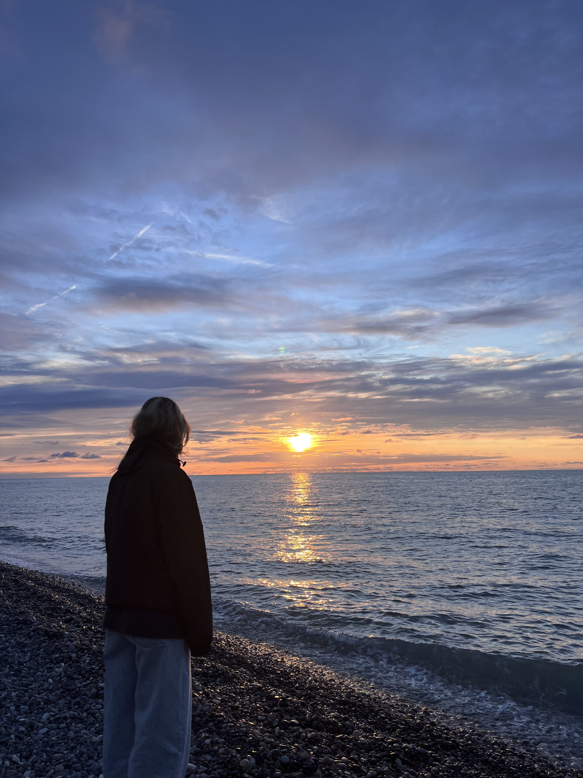 En kvinde ses fra ryggen, mens hun kigger ud over Det Sorte Hav på en stenstrand under en gylden solnedgang